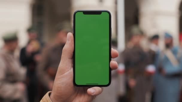 Closeup ενός επανδρώνει το χέρι που κρατά ένα κινητό τηλέφωνο με μια κάθετη πράσινη οθόνη στους δρόμους. Μεγάλη οθόνη. Στρατιωτική στρατιώτες στο παρασκήνιο — Αρχείο Βίντεο