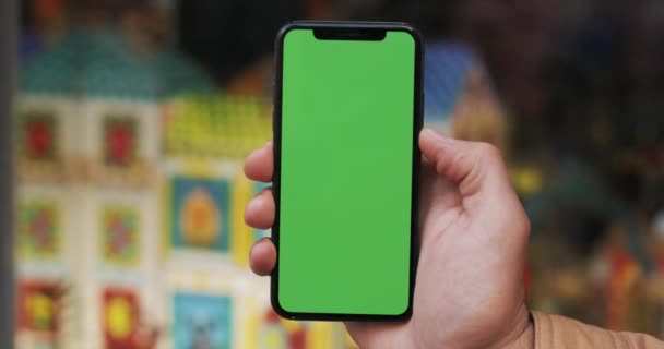 Closeup of a mans hand holding a mobile telephone with a vertical green screen on the streets. Childrens store in the background — Stock Video