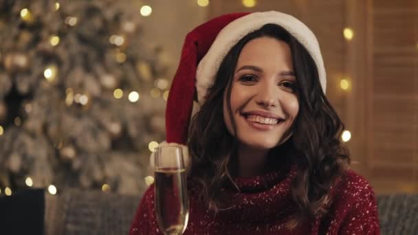 Привлекательная женщина со стаканом шампанского в Санта-Шляпе, смотрящая на камеру на фоне рождественской елки. Она поднимает стекло. Медленное движение — стоковое видео