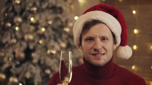 Hombre atractivo con una copa de champán en Santa Sombrero Mirando a la cámara en el fondo del árbol de Navidad. Levanta la copa. Movimiento lento — Vídeo de stock