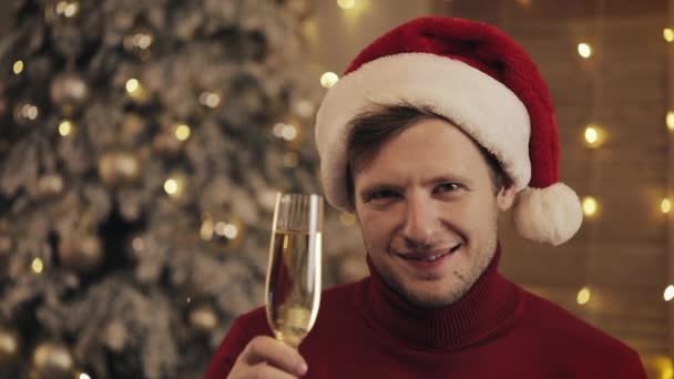 Hombre atractivo con una copa de champán en Santa Sombrero Mirando a la cámara en el fondo del árbol de Navidad. Levanta la copa. Movimiento lento — Vídeos de Stock