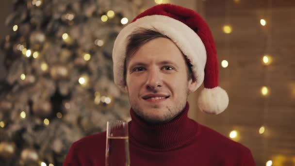 Hombre atractivo con una copa de champán en Santa Sombrero Mirando a la cámara en el fondo del árbol de Navidad. Levanta la copa. Movimiento lento — Vídeos de Stock