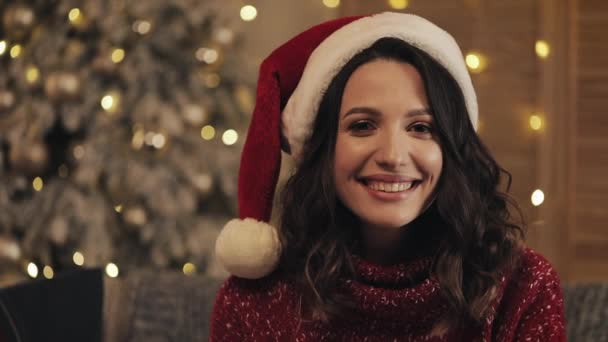 Porträt einer lächelnden attraktiven Frau mit Weihnachtsmütze, die auf dem Hintergrund des Weihnachtsbaums in die Kamera blickt. Zeitlupe — Stockvideo