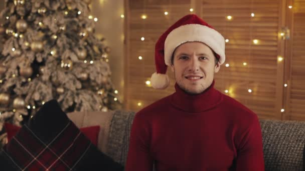 Retrato del hombre atractivo en Santa Sombrero Mirando a la cámara en el fondo del árbol de Navidad. Movimiento lento — Vídeos de Stock