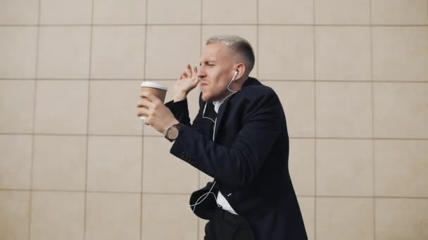 Счастливый молодой бизнесмен с кофе слушает музыку в наушниках на смартфоне и веселые танцы возле офисного здания. Смешные танцевальные движения. Медленное движение — стоковое видео