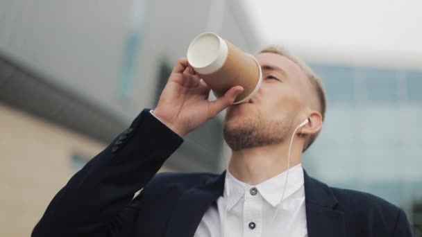 Feliz joven hombre de negocios con café escuchando música en auriculares en el teléfono inteligente y baile divertido cerca del edificio de oficinas. Divertidos movimientos de baile. Movimiento lento — Vídeos de Stock