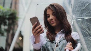 Yağışlı hava, gülümseyen, holding şemsiye, iletişim kavramı sokakta Smartphone kullanarak güzel genç iş kadını