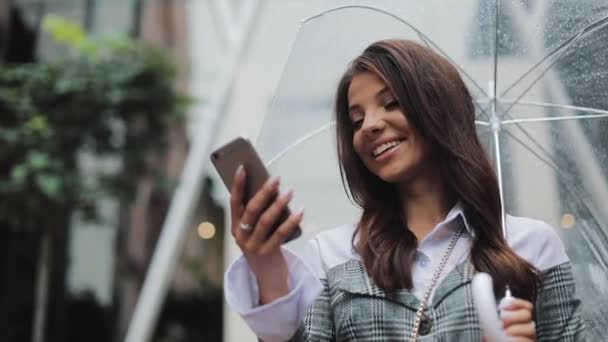 Hermosa joven mujer de negocios utilizando el teléfono inteligente en la calle en tiempo lluvioso, sonriendo, sosteniendo el paraguas, concepto de comunicación — Vídeos de Stock