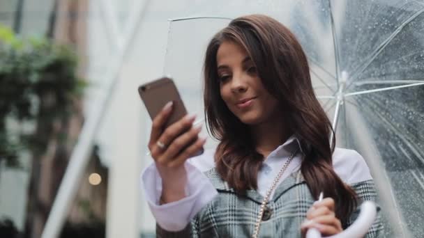 Yağışlı hava, gülümseyen, holding şemsiye, iletişim kavramı sokakta Smartphone kullanarak güzel genç iş kadını — Stok video