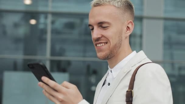 Ung affärsman med business-appen på smartphone stående i stad nära flygplats. Stilig ung affärsman kommunicerar på smartphone leende självsäker — Stockvideo