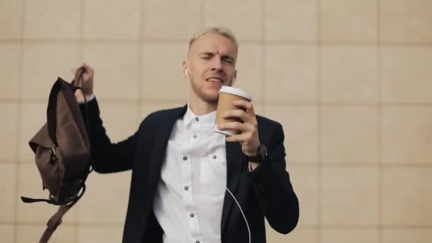 Joyeux jeune homme d'affaires avec café écoutant de la musique dans les écouteurs sur smartphone et danse drôle près de l'immeuble de bureaux. Des mouvements de danse drôles. Mouvement lent — Video