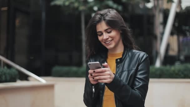Молодая счастливая женщина ходит по городской улице, пользуясь смартфоном. Медленное движение — стоковое видео