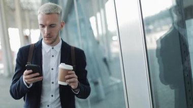 Genç çekici işadamı kahve tutarak ve telefon kullanarak sokakta yürüyor