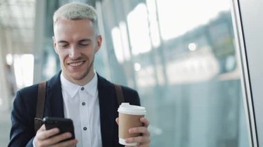 Genç çekici işadamı kahve tutarak ve telefon kullanarak sokakta yürüyor
