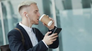 Genç çekici insan kahve tutarak ve telefon kullanarak sokakta yürüyor. İş Merkezi etrafında yürüyüş kulaklık müzik dinlediği