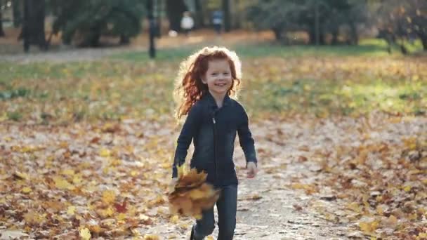 Маленькая симпатичная рыжая девушка, бегущая по осеннему парку и разбрасывающая листья — стоковое видео