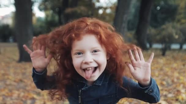 Крупным планом портрет милой рыжей девочки, стоящей в осеннем парке. Смешная девочка показывает гримасы и смеется — стоковое видео
