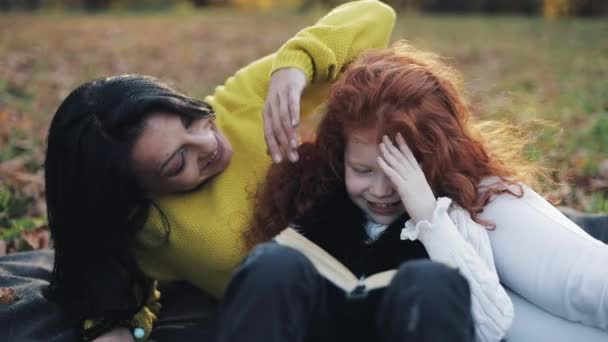 A mãe e a filha sentam-se na parte do Outono e lêem um livro. Eles falam bem. Família feliz na floresta de outono. Movimento lento — Vídeo de Stock