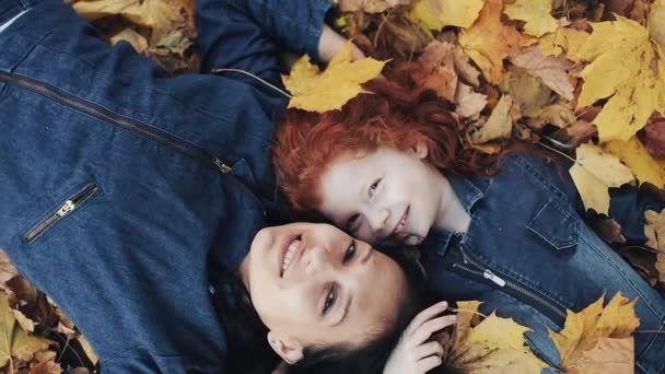 Eine schöne Mutter und ihre süße Tochter liegen auf einem gelben Blatt im herbstlichen Park. sie blicken in die Kamera, Draufsicht. Auf sie fallen gelbe Blätter. glückliche Familie hat Spaß. Zeitlupe — Stockvideo