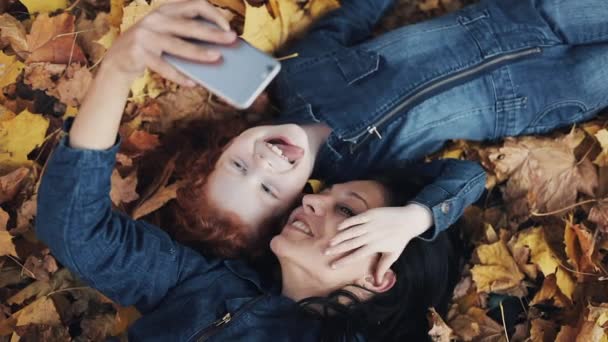 Μια όμορφη μητέρα και την κόρη της χαριτωμένο βρίσκονται σε ένα κίτρινο φύλλο στο φθινόπωρο park και κάνοντας μια selfie στο smartphone. Ευτυχισμένη οικογένεια διασκεδάζοντας. Το Top view, αργή κίνηση — Αρχείο Βίντεο