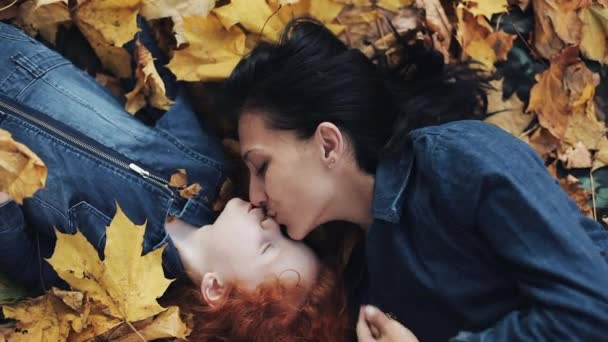 Una hermosa madre y su linda hija yacen sobre una hoja amarilla en el parque de otoño. Mamá besa a su hija. La familia feliz se está divirtiendo. Vista superior, cámara lenta — Vídeo de stock