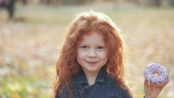 Маленькая милая рыжая девочка, стоящая в осеннем парке с пончиком в руке и смотрящая в камеру. Портрет, крупный план — стоковое видео