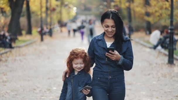 A beautiful mom and her cute little daughter walking in the autumn park. They using smartphones, laughing and have fun — Stock Video