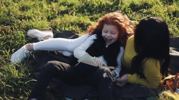 माँ और उसकी छोटी बेटी शरद ऋतु के हिस्से में बैठते हैं और एक किताब पढ़ते हैं। वे अच्छी तरह से बात करते हैं। शरद ऋतु जंगल में खुश परिवार। धीमी गति — स्टॉक वीडियो