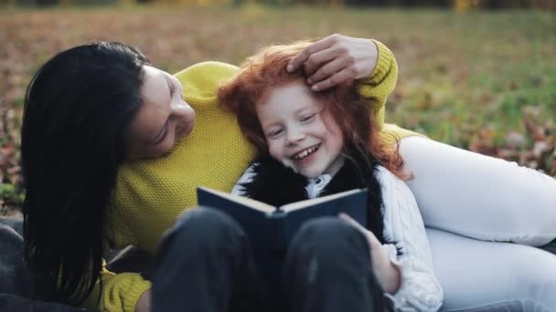 Mamá y su hija pequeña se sientan en la parte de otoño y leen un libro. Hablan muy bien. Familia feliz en el bosque de otoño. Movimiento lento — Vídeo de stock