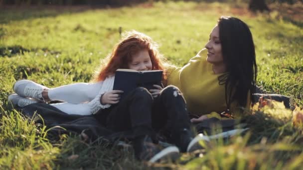 Mutter und kleine Tochter sitzen im Herbstteil und lesen ein Buch. Sie reden nett. glückliche Familie im Herbstwald. Zeitlupe — Stockvideo