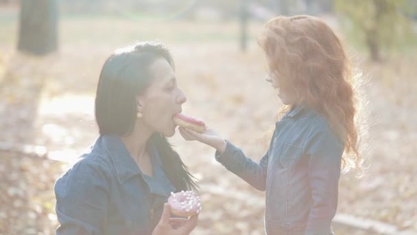 Красивая мама и ее рыжая дочь стоят в осеннем парке. Они едят пончики, кормят друг друга, веселятся — стоковое видео