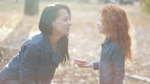 A beautiful mom and her pretty redhead daughter are standing in an autumn park. They eat donuts, feed each other, have fun — Stock Video