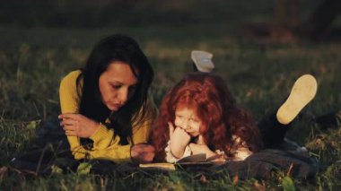 Güzel annem ve onun sevimli küçük kızı parkta çim üzerinde yalan ve bir kitap okuma. Güzel konuşurlar. Sonbahar ormanın içinde mutlu bir aile. Ağır çekim.