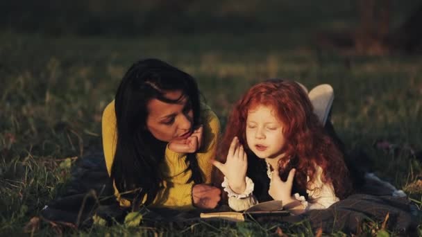 Belle mère et sa jolie petite fille allongées sur l'herbe dans le parc et lisant un livre. Ils parlent gentiment. Bonne famille dans la forêt d'automne. Mouvement lent — Video