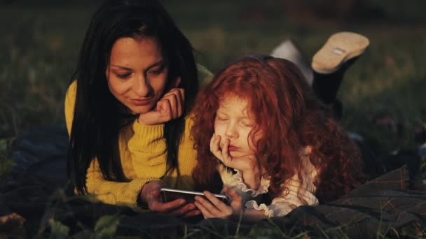 Die schöne Mutter und ihre süße kleine Tochter liegen auf dem Gras im Park und schauen zusammen Videos auf dem Smartphone. Urlaub, Kommunikation, Muttertagskonzept — Stockvideo