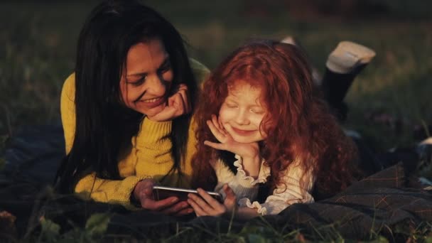 Hermosa madre y su linda hija pequeña tumbada en el césped en el parque y viendo video en el teléfono inteligente juntos. Vacaciones, comunicación, concepto del día de las madres — Vídeos de Stock