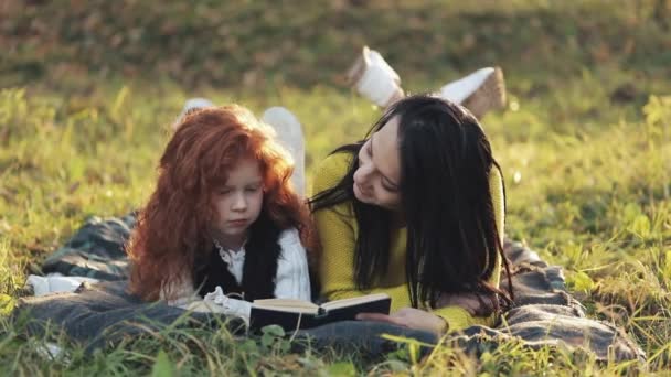 美丽的母亲和她可爱的小女儿躺在公园的草地上看书。他们说话很好。幸福的家庭在秋天的森林里。慢动作 — 图库视频影像