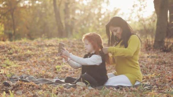 Bella giovane madre e la sua graziosa figlia rossa si divertono nel parco autunnale. La madre si prende cura di sua figlia, intreccia i capelli — Video Stock