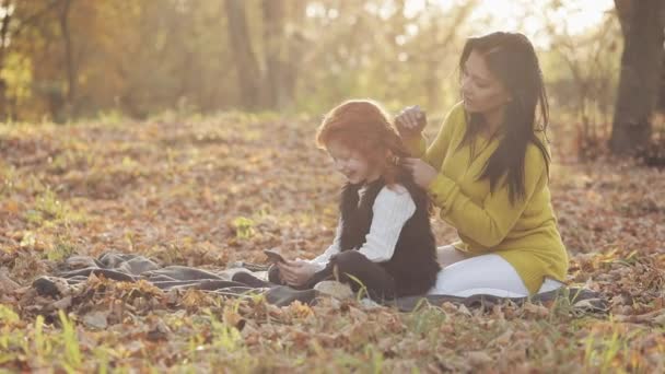 Güzel genç anne ve onun güzel kızıl saçlı kızı sonbahar parkta iyi eğlenceler. Annesi onun kızı ilgilenir, saçını örgü — Stok video