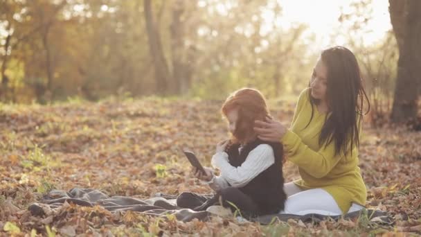 Молодая мама и ее симпатичная рыжая дочь веселятся в осеннем парке. Мать заботится о дочери, заплетает косички — стоковое видео