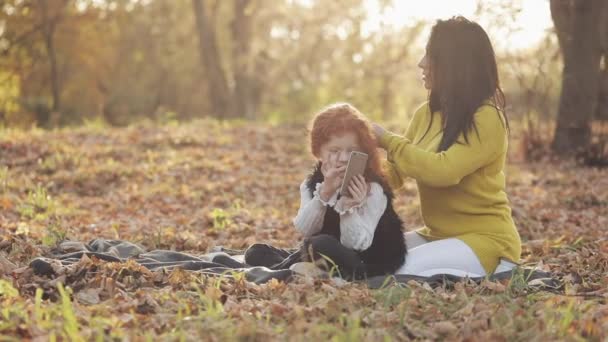 Hermosa madre joven y su linda hija pelirroja se divierten en el parque de otoño. La madre cuida de su hija, le trenza el pelo — Vídeos de Stock