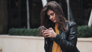 Genç ve güzel kadın kadın deri ceketli telefonunu dururken şehir merkezinde, etrafında görünüyor kullanır. Kentsel binalar arka plan