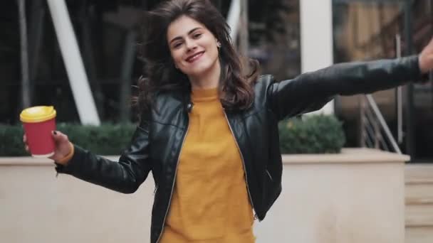 Mujer joven y feliz caminando cerca del centro de negocios y bailando con una taza de café en la mano. Hermosa chica mirando a la cámara. Buen humor, divertido, cámara lenta — Vídeos de Stock