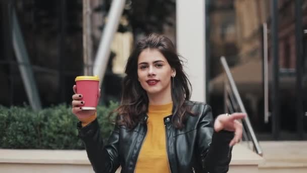 Mujer joven y feliz caminando cerca del centro de negocios y bailando con una taza de café en la mano. Hermosa chica mirando a la cámara. Buen humor, divertido, cámara lenta — Vídeos de Stock