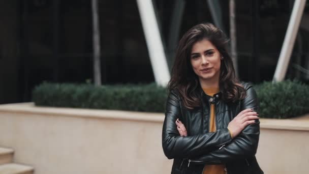 Portrait de la jeune femme séduisante debout près de l'immeuble de bureaux souriant à la caméra les mains croisées devant elle. Elle est vêtue d'un pull jaune chaud et d'une veste en cuir d'automne — Video