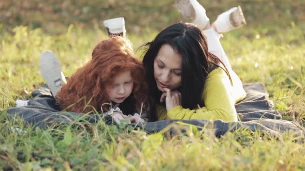 Belle mère et sa jolie petite fille allongées sur l'herbe dans le parc et lisant un livre. Ils parlent gentiment. Bonne famille dans la forêt d'automne. Mouvement lent — Video