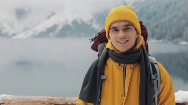 Portrait d'un jeune homme séduisant en vêtements d'hiver. Un randonneur portant des vêtements d'hiver jaunes regarde la caméra et sourit. Belles montagnes et fond de lac enneigé — Video