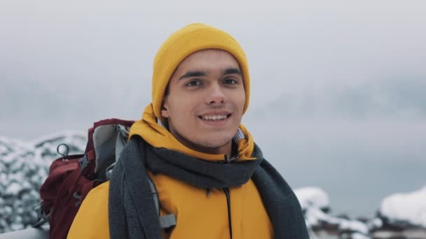 Портрет молодої привабливої людини в зимовому одязі. Похідний чоловік в жовтому зимовому одязі дивиться на камеру і посміхається. Красиві гори і засніжене озеро фону — стокове відео