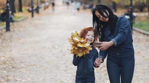 A beautiful mom and her cute little daughter walking in the autumn park. They making a selfie on a smartphone, laughing and have fun — Stock Video