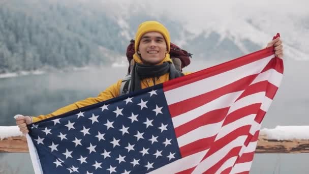 Podróżny mężczyznę trzymającego flagi America stojący w górach śniegiem jezioro przepiękne, miłe miejsce. Patrząc w kamerę i uśmiecha się. Turysta podróżny sobie w żółte zimowe ubrania — Wideo stockowe
